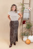 A woman modeling a beige short-sleeve mock neck top and a brown leopard print maxi skirt, standing in a studio decorated with a wooden ladder, flowers, and pumpkins.