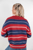 A woman viewed from behind wearing a red and navy blue striped knit sweater with thin white horizontal stripes, paired with blue jeans against a light gray studio background.