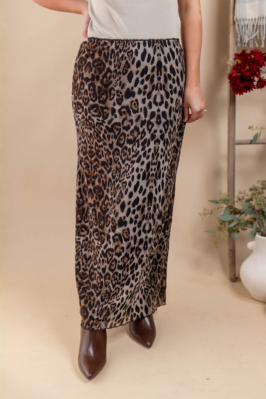 A woman models a leopard-print maxi skirt with brown pointed-toe boots, standing in a styled indoor setting with a beige backdrop, decorative flowers, and a white vase.