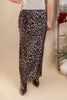 A woman models a leopard-print maxi skirt with brown pointed-toe boots, standing in a styled indoor setting with a beige backdrop, decorative flowers, and a white vase.