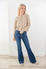 A woman modeling a beige pointelle cable-knit cropped cardigan sweater with buttons, paired with blue flare jeans, standing in a bright room with white walls and light wood flooring.