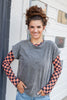 A woman wearing a black gray mineral washed short-sleeve top with a Halloween long sleeve orange and black checkered under, standing outdoors against a white building wall.