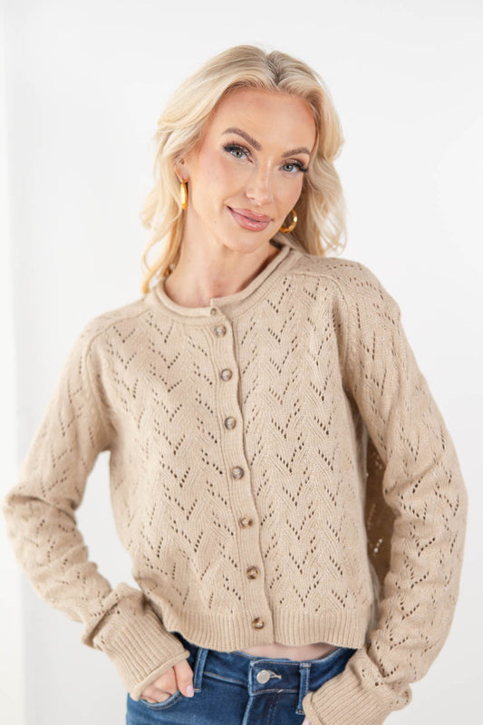 A blonde woman wearing a beige pointelle cable-knit button-up cardigan sweater with blue jeans, photographed against a clean white studio background.
