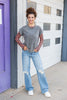 A woman stands outdoors in front of a white garage door and purple door frame, wearing a gray short-sleeve top with orange and black checkered long sleeves, light blue distressed jeans, and white sneakers.