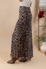 A woman wearing a leopard print maxi skirt with brown pointed-toe heels, standing against a beige studio backdrop with decorative plants and flowers on the side.