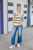 A woman smiling in front of a white garage, wearing a short-sleeve knit polo shirt with horizontal olive green and white stripes, a white collar, and a split V-neck with wide leg blue jeans.