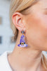 A close-up of a model's ear wearing a dangling earring shaped like a purple witch hat with a small spider charm at the top.