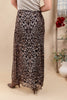 A woman wearing a leopard-print maxi skirt with brown heeled boots, shown from the back in a styled indoor setting with a beige wall and decorative plants.