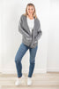 A woman modeling a gray open-front knit cardigan with white geometric patterns on the sleeves, paired with a white t-shirt, blue skinny jeans, and white sneakers, standing against a white wall and light wood floor.