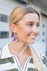 A smiling blonde woman in profile wearing a small purple witch hat earring with gold trim, photographed outdoors against a softly blurred building background.