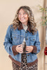 A woman wearing a blue denim jacket with brown suede-like collar, cuffs, and hem, layered over a beige top and leopard print skirt, posing against a warm beige background.