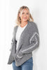A woman wearing a chunky gray knit cardigan with white textured blanket stitch detail on the sleeves, styled over a white t-shirt and blue jeans against a plain white background.