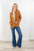 A woman modeling a warm camel-colored cardigan with front pockets over a white polka-dot top and blue flared jeans, standing in a bright room with white walls and light wood flooring.