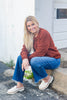 A smiling blonde woman sits on a stone curb outdoors, wearing a rust-colored knit henley sweater, blue jeans, and cream slip-on loafers.