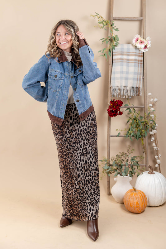 A woman models a light blue denim jacket with brown suede collar and cuffs, paired with a brown and black leopard print maxi skirt and brown leather boots, set against a warm beige studio backdrop with decorative autumn props.
