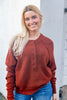 A smiling blonde woman wearing a rust-brown long-sleeve henley sweater and blue jeans, standing against a light gray wall.