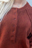 Close-up of a person wearing a rust-brown knit sweater with a buttoned henley-style neckline. The sweater has a soft, textured knit and raglan sleeves, shown against a light background with the model's blonde hair visible on the left.