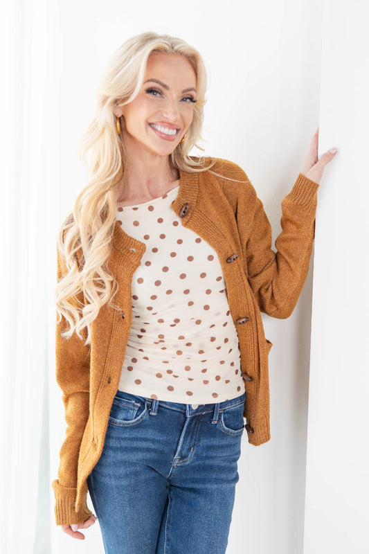 A smiling blonde woman leans against a white wall wearing a camel brown button-up cardigan over a cream polka-dot top and blue jeans.