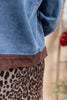 Close-up of a person wearing a light blue denim jacket with a brown suede-like hem, paired with a leopard-print skirt, against a softly blurred beige background.