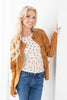 A smiling blonde woman leans against a white wall wearing a camel brown button-up cardigan over a cream polka-dot top and blue jeans.