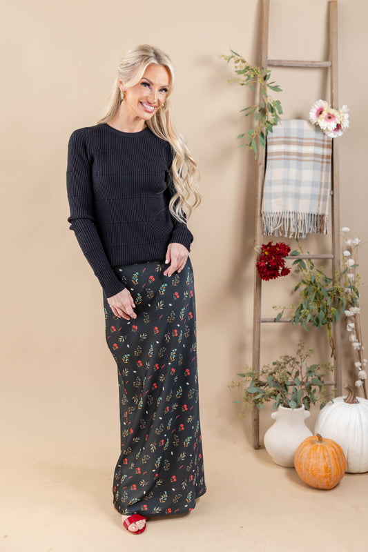 A woman stands in a studio wearing a black ribbed long-sleeve sweater and a black maxi skirt with a red and yellow floral print. She is posed next to a rustic wooden ladder decorated with a plaid throw blanket, white vases with greenery, and pumpkins against a solid beige background.