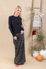A woman stands in a studio wearing a black ribbed long-sleeve sweater and a black maxi skirt with a red and yellow floral print. She is posed next to a rustic wooden ladder decorated with a plaid throw blanket, white vases with greenery, and pumpkins against a solid beige background.