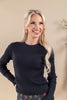 A smiling blonde woman wearing a black ribbed knit sweater and dark floral pants, standing against a warm beige background.