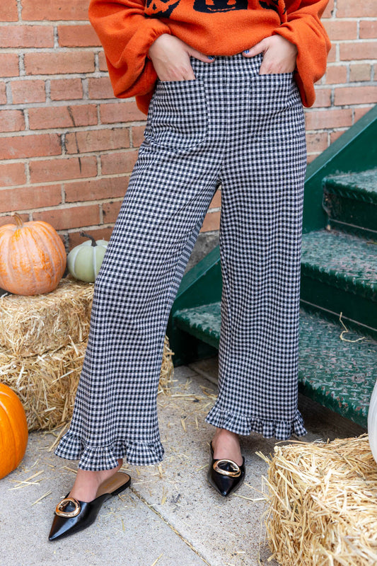 A person wearing black and white gingham wide-leg pants with ruffled hems, paired with an orange sweatshirt and black pointed flats, standing outdoors near hay bales, pumpkins, and green steps.
