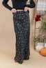 A woman wearing a long black maxi skirt with a small multicolor floral print, paired with a dark ribbed sweater. The photo is taken in a styled indoor setting with a beige backdrop, a wooden ladder, and autumn decorations like a pumpkin and potted plants.