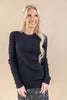 A smiling blonde woman wearing a black ribbed knit sweater and a dark floral skirt, standing against a solid beige studio background.