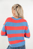 A woman viewed from behind wearing a knit sweater with horizontal coral red and medium blue stripes, paired with blue jeans against a plain white background.