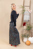A woman posing in a black ribbed long-sleeve sweater and a black maxi skirt with a red and yellow floral pattern, standing in a studio decorated with a wooden ladder, pumpkins, and potted plants.