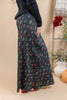 A woman wearing a long black maxi skirt with a colorful floral and leaf print, paired with a dark ribbed sweater. The photo is taken in a warm, neutral-toned indoor setting with decorative plants and a pumpkin in the background.