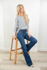 A woman with blonde hair sits on a wooden stool, wearing a gray long-sleeve top and dark blue flare jeans in a bright room with white walls and light wood flooring.