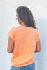 A woman viewed from behind wearing a short-sleeve, crew-neck t-shirt in a washed orange color, paired with blue jeans, standing against a textured white wall.
