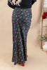 A woman wearing a black maxi skirt with a small multicolor floral print, paired with a black ribbed sweater and red shoes, shown from the back against a beige studio backdrop.