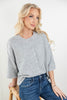 A blonde woman wearing a light gray heather knit cardigan with a button-down front and blue jeans, seated on a wooden stool against a plain white background.