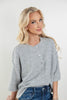 A blonde woman wearing a light gray heathered knit button-front top with elbow-length sleeves and blue jeans, photographed against a clean light gray studio background.