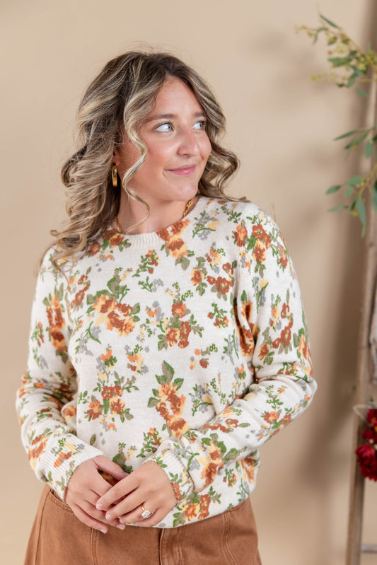 A woman wearing a cream-colored crewneck sweater with an orange and green floral pattern, paired with brown pants, standing against a warm beige background.