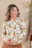 A woman wearing a cream-colored crewneck sweater with an orange and green floral pattern, paired with brown pants, standing against a warm beige background.
