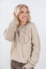 A smiling blonde woman wearing a beige crewneck sweater with a brown line art dog graphic on the front, paired with brown jeans, against a plain light gray background.