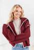 A woman wearing a chunky ribbed knit cardigan in deep burgundy with white scalloped trim, layered over a beige turtleneck and blue jeans.