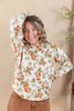 A woman smiling and posing in a cream-colored long-sleeve sweatshirt with an orange and green floral pattern, paired with brown pants, set against a warm beige background.