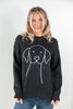 A woman wearing a black crewneck sweater with a white line art graphic of a dog on the front, paired with blue jeans, photographed against a light gray studio background.