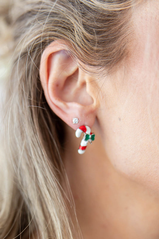 A close-up of a woman's ear wearing a small silver stud earring with a dangling candy cane charm in red, white, and green enamel.