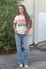 A woman stands outdoors wearing a cream graphic t-shirt with colorful text that reads 'Pumpkin S'mores Hayrides Bonfires', paired with light-wash blue jeans and white boots.