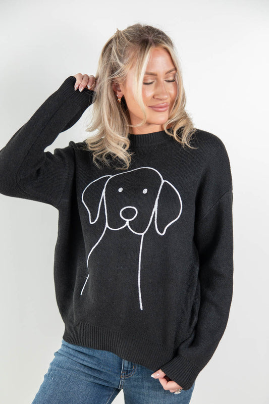 A woman wearing a black knit sweater with a white line art graphic of a dog on the front, paired with blue jeans, against a plain light gray background.