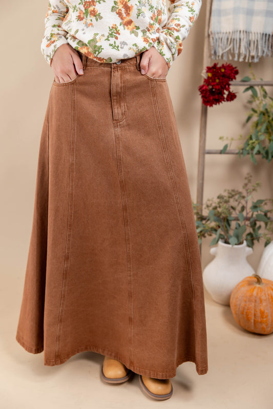 A person wearing a long, flared brown corduroy skirt with a button fly and front pockets, paired with a floral long-sleeve top and tan boots, standing in a styled indoor setting with a pumpkin and potted plants.