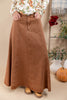 A person wearing a long, flared brown corduroy skirt with a button fly and front pockets, paired with a floral long-sleeve top and tan boots, standing in a styled indoor setting with a pumpkin and potted plants.