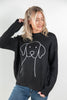 A woman wearing a black knit sweater with a white line art graphic of a dog on the front, paired with blue jeans, against a plain light gray background.